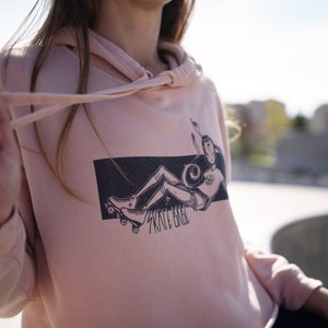 Peut inclure: Un sweat &agrave; capuche rose avec un imprim&eacute; graphique noir et blanc d'une personne sur des patins &agrave; roulettes. Le graphique comprend le texte "Skate Hard".