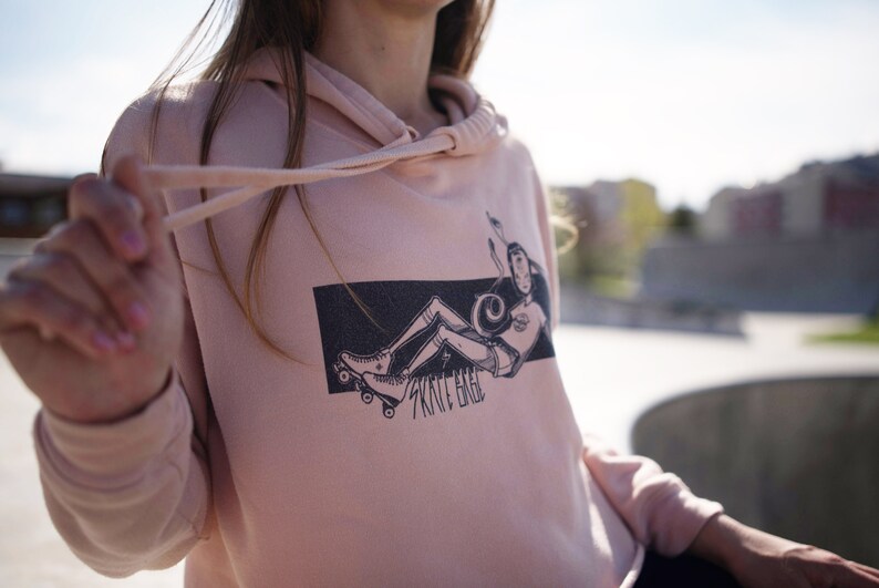 Peut inclure: Un sweat &agrave; capuche rose avec un imprim&eacute; graphique noir et blanc d'une personne sur des patins &agrave; roulettes. Le graphique comprend le texte "Skate Hard".