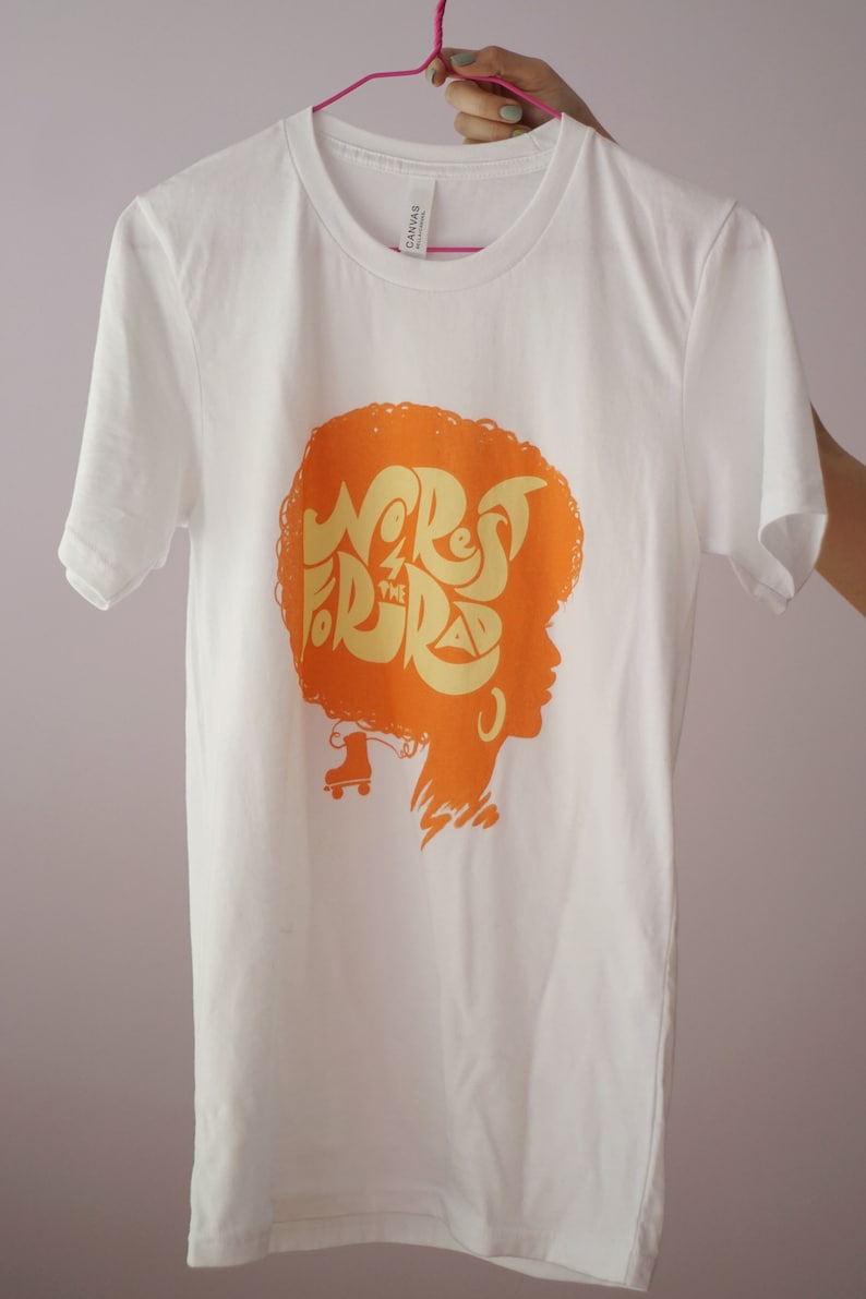 May include: White t-shirt featuring an orange graphic of a woman's silhouette with an afro hairstyle. The graphic reads "Wrest for the Rad" in a yellow font.