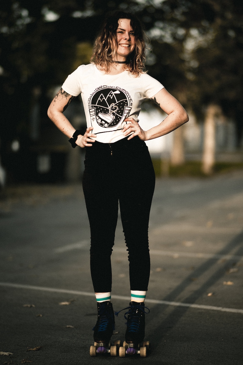 May include: A woman wearing a white t-shirt with a black and white graphic of a roller skate and mountains. The shirt says "Roll Grow Empower". She is wearing black pants and teal and white striped socks. She is standing on roller skates with purple wheels.