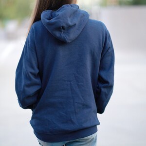 Puede incluir: Una persona lleva una sudadera con capucha azul marino con la capucha puesta y pantalones cortos de mezclilla. Lleva calcetines blancos con rayas azules y amarillas.