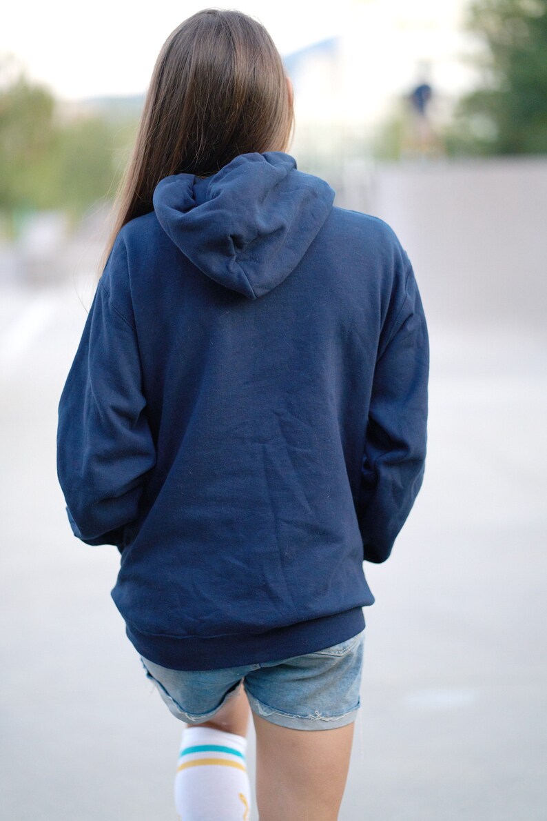 Puede incluir: Una persona lleva una sudadera con capucha azul marino con la capucha puesta y pantalones cortos de mezclilla. Lleva calcetines blancos con rayas azules y amarillas.