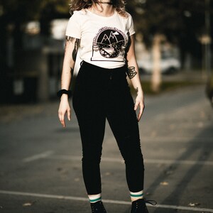 May include: A woman wearing a white t-shirt with a black and white graphic of mountains and the text "Roll, Grow, Empower" is roller skating on a black asphalt surface. She is wearing black pants, white socks with teal stripes, and black roller skates with teal wheels.