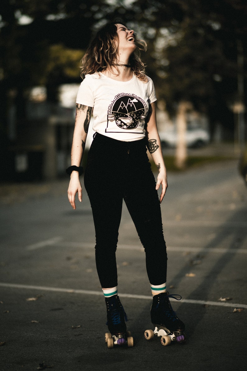 May include: A woman wearing a white t-shirt with a black and white graphic of mountains and the text "Roll, Grow, Empower" is roller skating on a black asphalt surface. She is wearing black pants, white socks with teal stripes, and black roller skates with teal wheels.