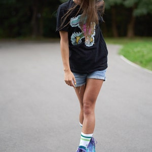 Puede incluir: Una mujer con una camiseta negra con un estampado gr&aacute;fico colorido y pantalones cortos de mezclilla est&aacute; patinando sobre ruedas en un camino pavimentado. Lleva patines azules y morados con calcetines blancos.