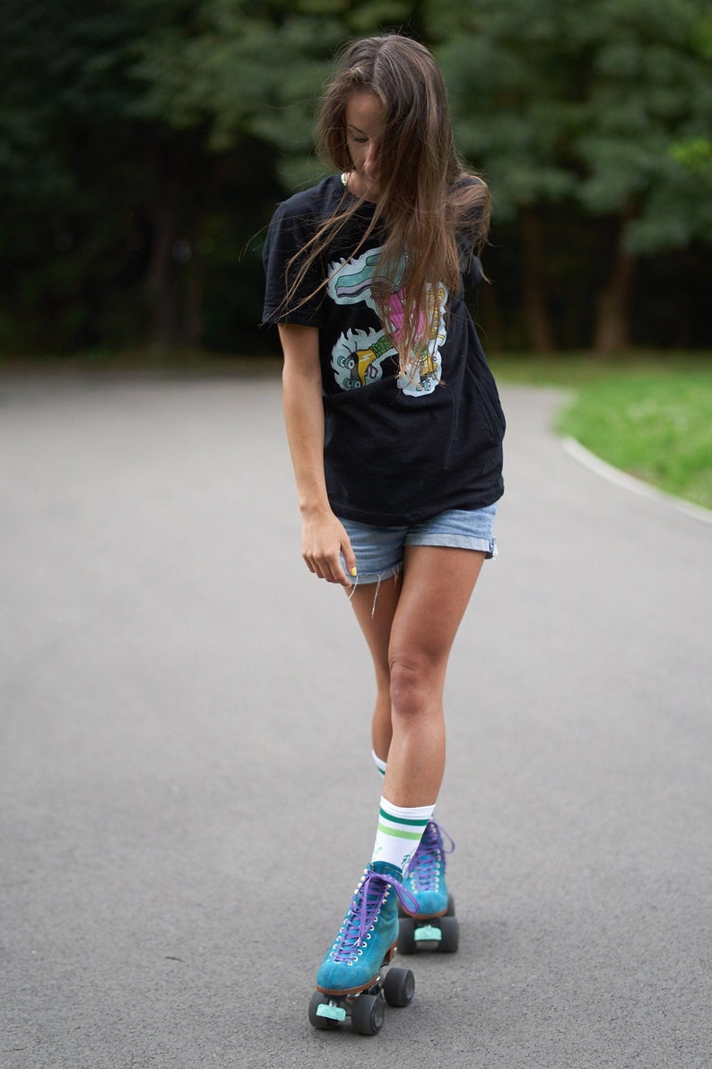 Puede incluir: Una mujer con una camiseta negra con un estampado gr&aacute;fico colorido y pantalones cortos de mezclilla est&aacute; patinando sobre ruedas en un camino pavimentado. Lleva patines azules y morados con calcetines blancos.