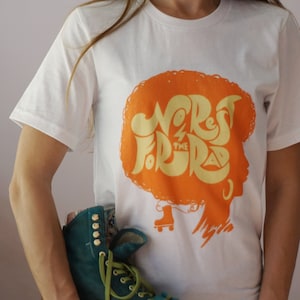 May include: White t-shirt featuring an orange graphic of a woman's silhouette with the text "No Rest For The Rad" in yellow lettering. The woman is wearing roller skates.