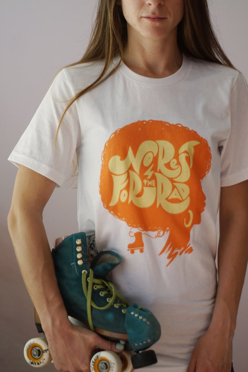 May include: White t-shirt featuring an orange graphic of a woman's silhouette with the text "No Rest For The Rad" in yellow lettering. The woman is wearing roller skates.