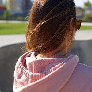 Peut inclure: Une femme avec de longs cheveux bruns portant un sweat &agrave; capuche rose. La capuche est tir&eacute;e sur sa t&ecirc;te. Elle porte des lunettes de soleil et regarde loin de l'appareil photo.
