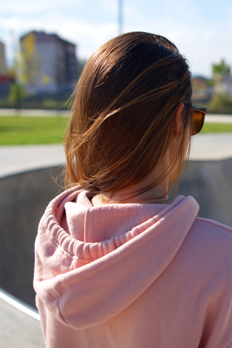 Peut inclure: Une femme avec de longs cheveux bruns portant un sweat &agrave; capuche rose. La capuche est tir&eacute;e sur sa t&ecirc;te. Elle porte des lunettes de soleil et regarde loin de l'appareil photo.