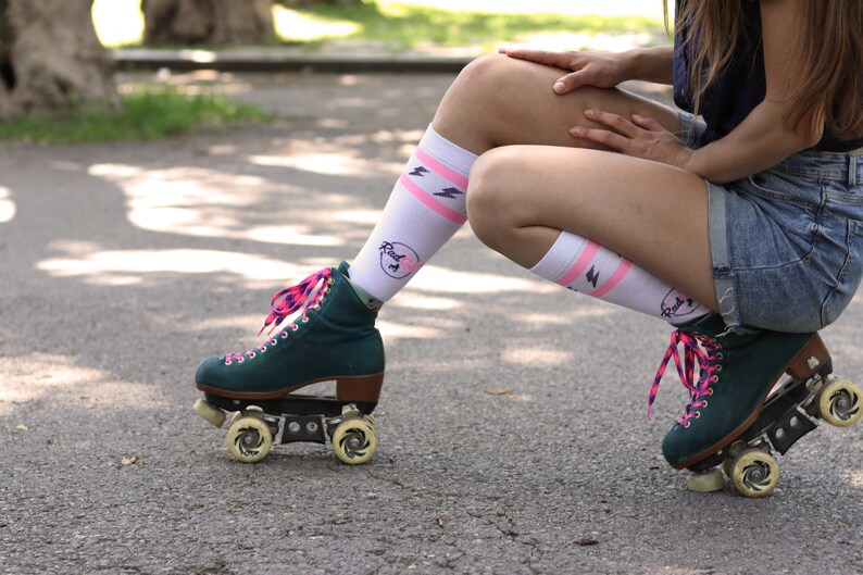 K&ouml;nnte beinhalten: Eine Person tr&auml;gt wei&szlig;e Socken mit rosa Streifen und einem Blitz-Design sowie gr&uuml;ne Rollschuhe mit rosa Schn&uuml;rsenkeln. Die Socken haben einen rosa Kreis mit dem Text "Roll 1" darauf.