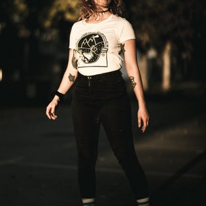 May include: A woman wearing a white t-shirt with a black and white graphic of mountains and a compass. She is wearing black jeans and roller skates. The background is blurred and dark.