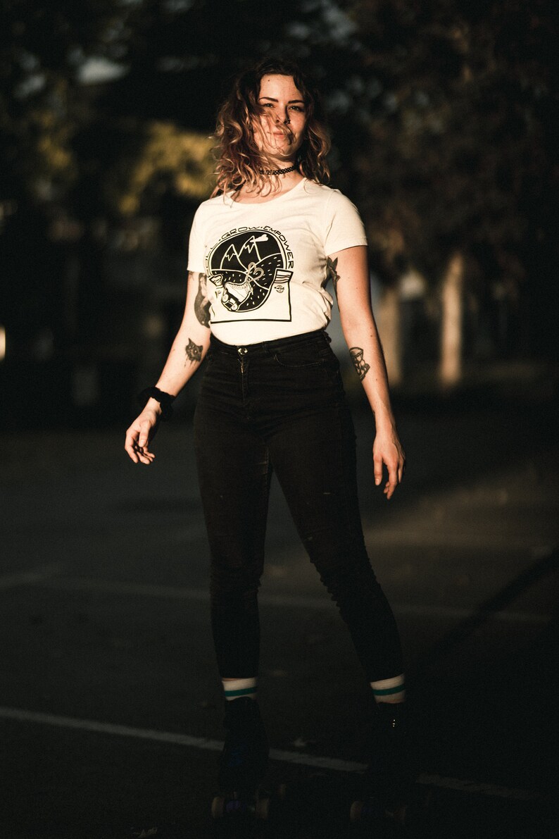 May include: A woman wearing a white t-shirt with a black and white graphic of mountains and a compass. She is wearing black jeans and roller skates. The background is blurred and dark.