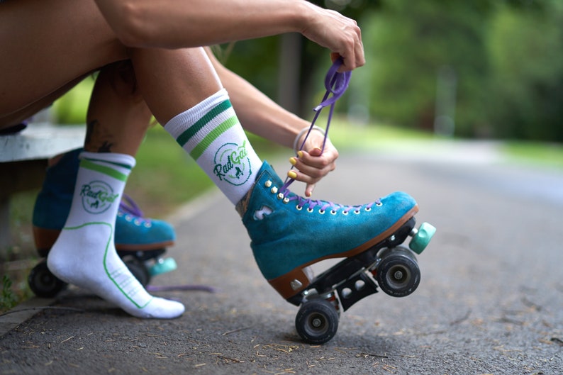 Puede incluir: Una persona lleva calcetines blancos con rayas verdes y el texto "Rad Gal" en verde. Est&aacute; atando los cordones de un pat&iacute;n de color azul verdoso con cordones morados.