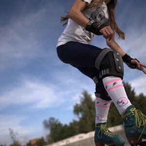 K&ouml;nnte beinhalten: Eine Frau tr&auml;gt wei&szlig;e Socken mit rosa Blitzen und dem Schriftzug "Rad Girl" beim Rollschuhlaufen im Freien. Sie tr&auml;gt schwarze Knieschoner und schwarze Handgelenksch&uuml;tzer.