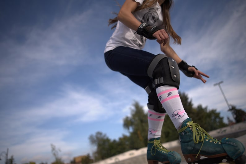 K&ouml;nnte beinhalten: Eine Frau tr&auml;gt wei&szlig;e Socken mit rosa Blitzen und dem Schriftzug "Rad Girl" beim Rollschuhlaufen im Freien. Sie tr&auml;gt schwarze Knieschoner und schwarze Handgelenksch&uuml;tzer.