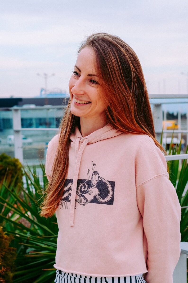 Peut inclure: Une femme portant un sweat &agrave; capuche rose court avec un imprim&eacute; graphique noir et blanc d'un personnage de dessin anim&eacute;. Le sweat &agrave; capuche a une capuche &agrave; cordon et une coupe d&eacute;contract&eacute;e. Elle sourit et regarde vers la droite.