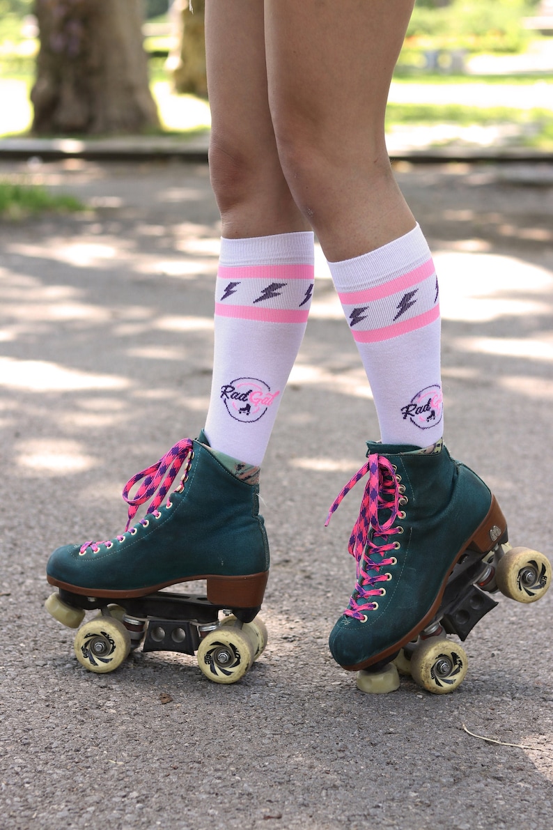 K&ouml;nnte beinhalten: Ein Paar gr&uuml;ne Rollschuhe mit rosa und violetten Schn&uuml;rsenkeln. Die Schuhe haben wei&szlig;e Socken mit rosa Streifen und Blitzmustern. Die Socken haben den Text "Rad Girl" mit einer Rollschuhgrafik.