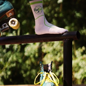 Puede incluir: Una persona lleva calcetines blancos con rayas verdes y blancas y el texto "Rad Gal" en verde. La persona est&aacute; de pie sobre una barandilla de metal negro con un pie. Un pat&iacute;n de ruedas azul verdoso con cordones amarillos est&aacute; en el suelo debajo de la barandilla.