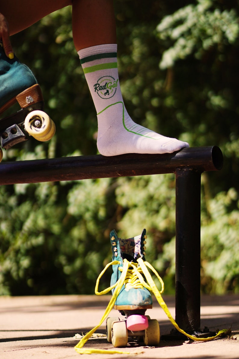 Puede incluir: Una persona lleva calcetines blancos con rayas verdes y blancas y el texto "Rad Gal" en verde. La persona est&aacute; de pie sobre una barandilla de metal negro con un pie. Un pat&iacute;n de ruedas azul verdoso con cordones amarillos est&aacute; en el suelo debajo de la barandilla.
