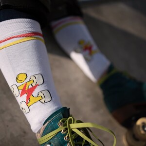 Op de afbeelding: Witte kniehoge sokken met rode en gele strepen aan de bovenkant. De sokken hebben een afbeelding van een persoon die rolschaatst met een bliksem. De persoon draagt ​​turkooise rollerskates met groene veters.