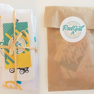 Puede incluir: Un par de calcetines blancos con un patr&oacute;n amarillo y azul est&aacute;n envueltos en papel marr&oacute;n y atados con una cuerda. Una peque&ntilde;a tarjeta con un dise&ntilde;o de patines y el texto "Rad Gal Roller Skate Accessories" est&aacute; unida al paquete.