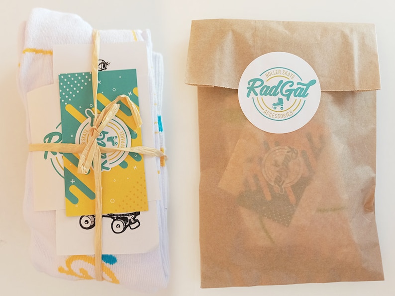 Puede incluir: Un par de calcetines blancos con un patr&oacute;n amarillo y azul est&aacute;n envueltos en papel marr&oacute;n y atados con una cuerda. Una peque&ntilde;a tarjeta con un dise&ntilde;o de patines y el texto "Rad Gal Roller Skate Accessories" est&aacute; unida al paquete.