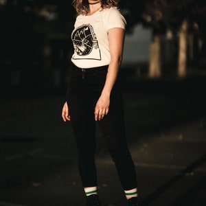 May include: A woman wearing a white t-shirt with a black graphic print and black pants is roller skating. She is wearing green and white striped socks and black roller skates.