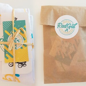 Puede incluir: Un par de calcetines blancos con un patr&oacute;n amarillo y azul est&aacute;n envueltos en papel marr&oacute;n y atados con una cuerda. Una tarjeta con un dise&ntilde;o amarillo y verde y el texto "Rad Gal Roller Skate Accessories" est&aacute; unida al paquete.