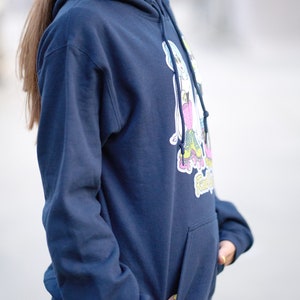 Puede incluir: Una sudadera con capucha azul marino con un gr&aacute;fico de dos personas patinando. El gr&aacute;fico es en rosa, azul, amarillo y verde. La sudadera tiene un bolsillo canguro.