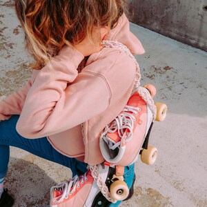 Peut inclure: Une personne portant un jean bleu et un sweat-shirt rose tient une paire de rollers roses avec des lacets blancs et des roues blanches.