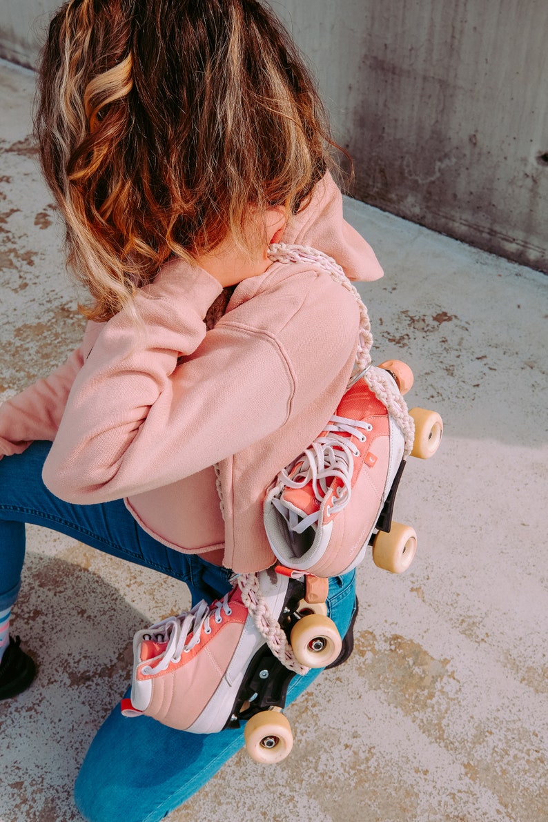 Peut inclure: Une personne portant un jean bleu et un sweat-shirt rose tient une paire de rollers roses avec des lacets blancs et des roues blanches.