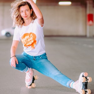 May include: A woman wearing a white t-shirt with an orange graphic and blue jeans is roller skating. She is wearing pink roller skates with white wheels.