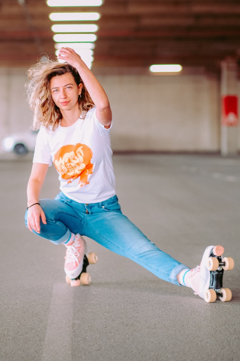 May include: A woman wearing a white t-shirt with an orange graphic and blue jeans is roller skating. She is wearing pink roller skates with white wheels.