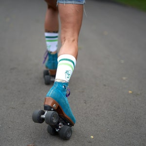 Puede incluir: Una persona que lleva pantalones cortos de mezclilla azul y calcetines blancos con rayas verdes y un logotipo que dice "Rad" est&aacute; patinando sobre ruedas en un camino pavimentado. La persona lleva patines azules con ruedas marrones.