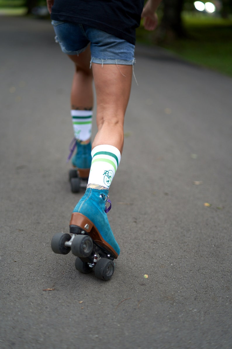 Puede incluir: Una persona que lleva pantalones cortos de mezclilla azul y calcetines blancos con rayas verdes y un logotipo que dice "Rad" est&aacute; patinando sobre ruedas en un camino pavimentado. La persona lleva patines azules con ruedas marrones.