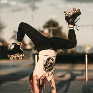 May include: A person wearing black jeans and a white t-shirt with a graphic design of a roller skate. They are doing a handstand with their roller skates in the air.