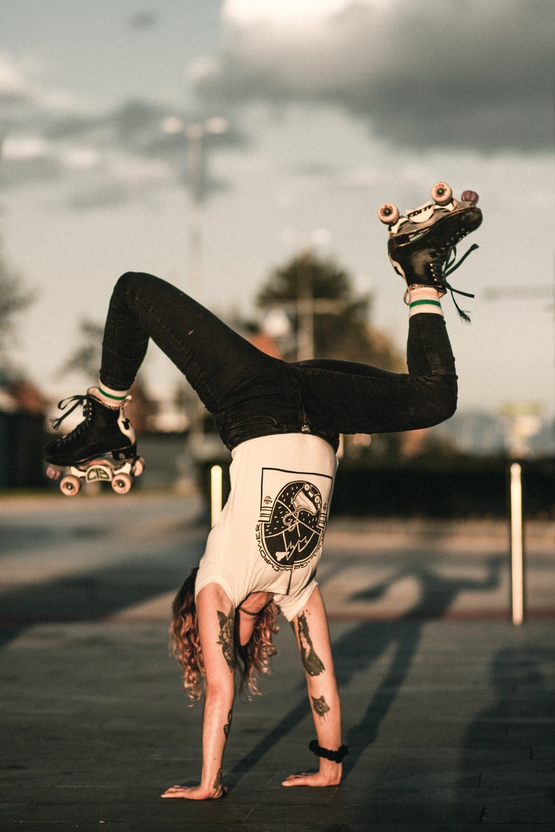 May include: A person wearing black jeans and a white t-shirt with a graphic design of a roller skate. They are doing a handstand with their roller skates in the air.
