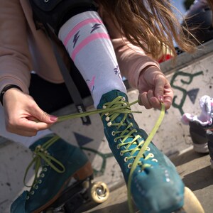 K&ouml;nnte beinhalten: Eine Person tr&auml;gt t&uuml;rkisfarbene Rollschuhe mit gr&uuml;nen Schn&uuml;rsenkeln und bindet die Schn&uuml;rsenkel. Die Rollschuhe haben wei&szlig;e Rollen und schwarze Rahmen.