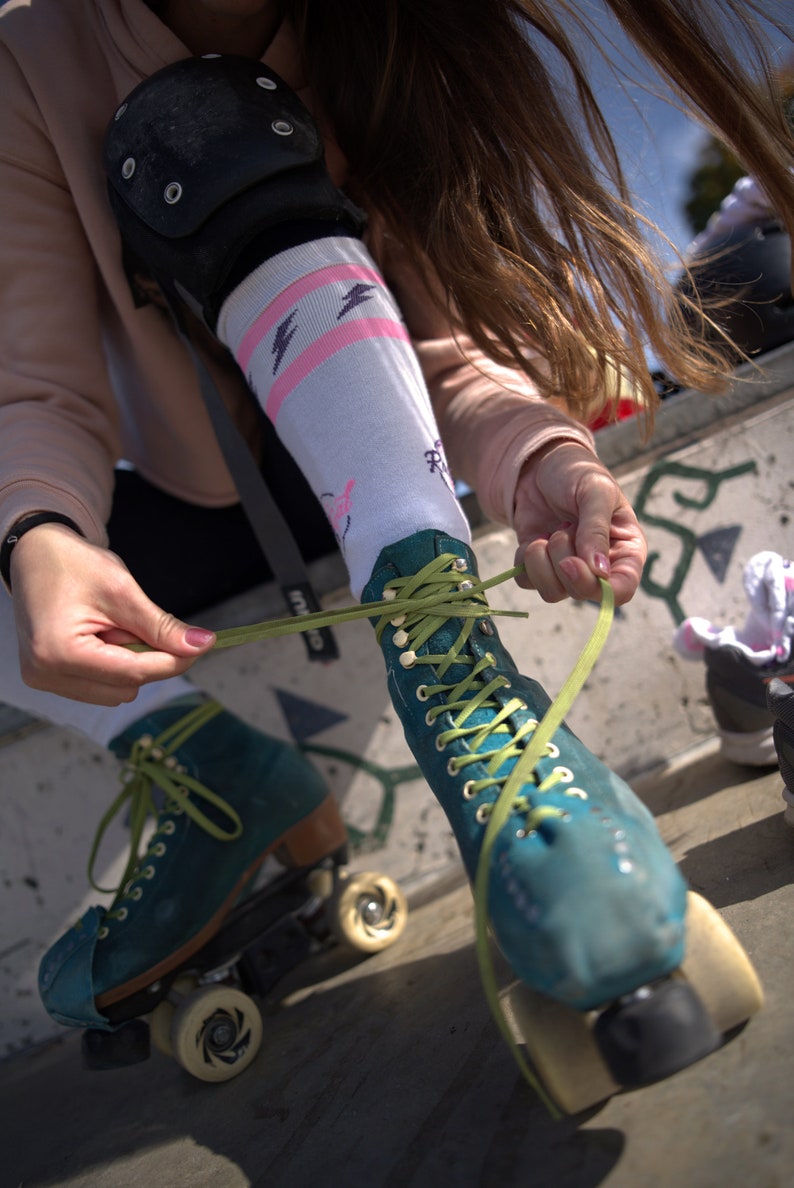 K&ouml;nnte beinhalten: Eine Person tr&auml;gt t&uuml;rkisfarbene Rollschuhe mit gr&uuml;nen Schn&uuml;rsenkeln und bindet die Schn&uuml;rsenkel. Die Rollschuhe haben wei&szlig;e Rollen und schwarze Rahmen.