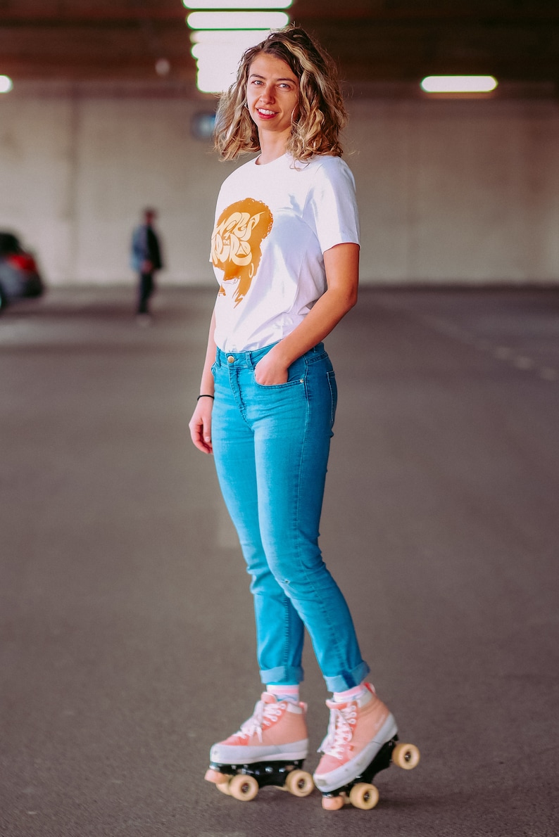 May include: A woman wearing a white t-shirt with a gold graphic and blue jeans is roller skating. She is wearing pink roller skates with white wheels.