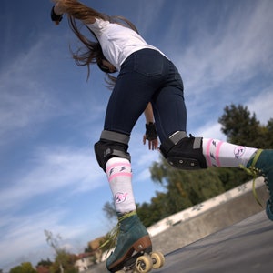 K&ouml;nnte beinhalten: Eine Frau mit Rollschuhen, Knieschonern und Socken mit pinkfarbenen und wei&szlig;en Blitzen f&auml;hrt auf einer Rampe. Der Himmel ist blau mit wei&szlig;en Wolken.