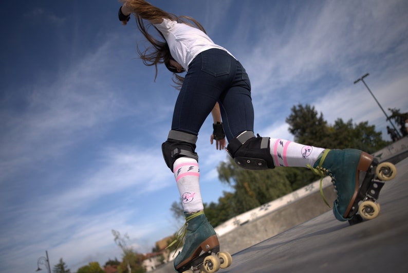 K&ouml;nnte beinhalten: Eine Frau mit Rollschuhen, Knieschonern und Socken mit pinkfarbenen und wei&szlig;en Blitzen f&auml;hrt auf einer Rampe. Der Himmel ist blau mit wei&szlig;en Wolken.