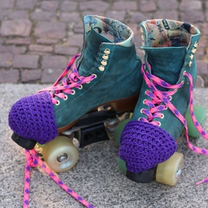 Puede incluir: Un par de patines de ruedas de color turquesa con cubrededos de ganchillo morado. Los patines tienen cordones de color rosa y morado y están sobre una superficie de ladrillo.