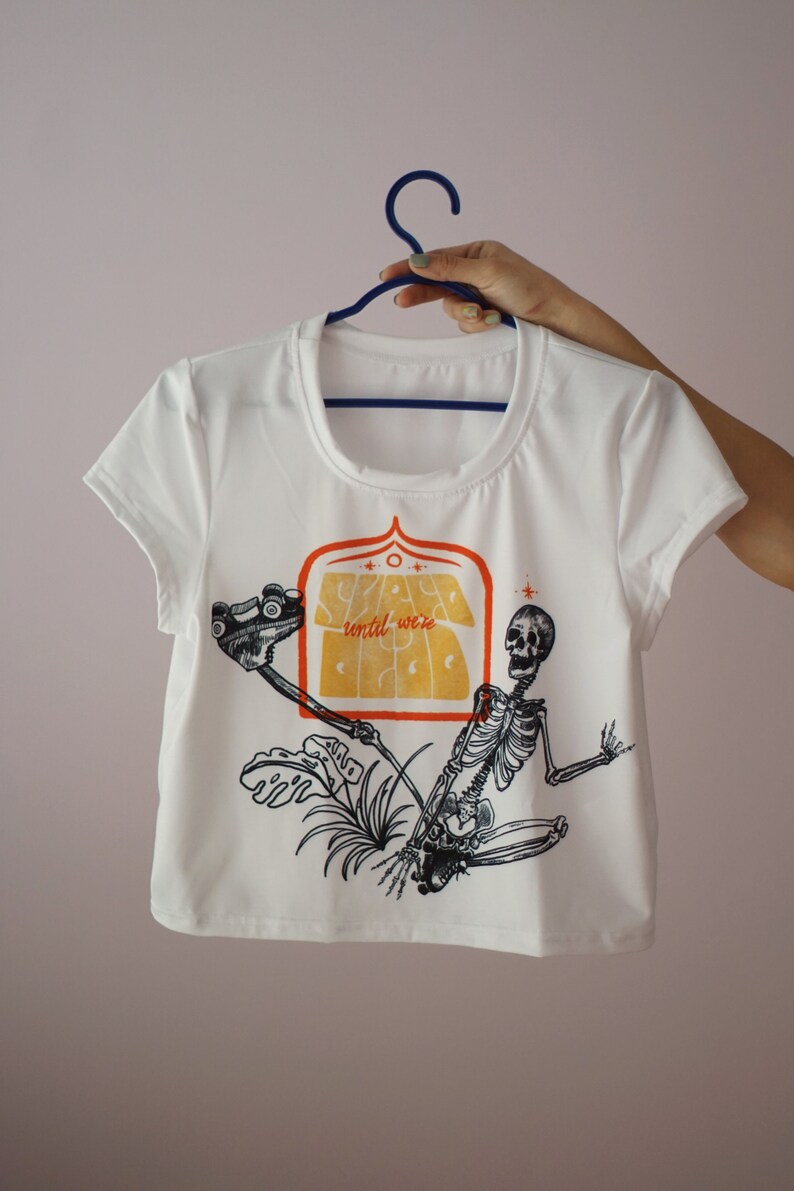 May include: White t-shirt with a graphic print of a skeleton holding a roller skate and a sign that says "Stay until were sick".