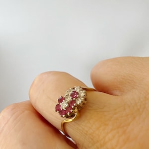 May include: A gold ring featuring a cluster of red gemstones and clear gemstones. The ring's design is floral, with the gemstones arranged to resemble a flower. The band is a simple, elegant gold. The ring is shown on a finger.