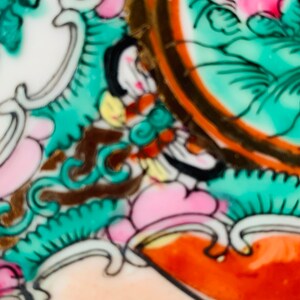 May include: Close-up of a ceramic plate with a colorful floral design. The design features pink, green, orange, and gold accents. The plate is decorated with intricate details and a swirling pattern.