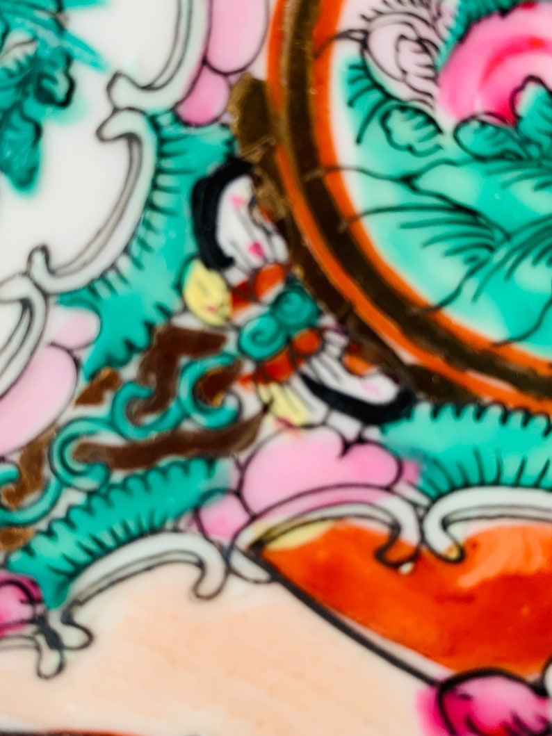 May include: Close-up of a ceramic plate with a colorful floral design. The design features pink, green, orange, and gold accents. The plate is decorated with intricate details and a swirling pattern.