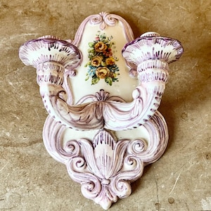 May include: A white ceramic wall sconce with two candle holders. The sconce is decorated with purple and pink floral designs and has a floral motif in the centre.