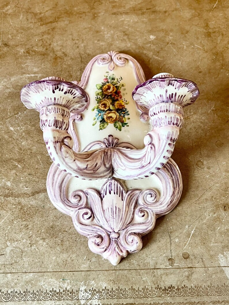 May include: A white ceramic wall sconce with two candle holders. The sconce is decorated with purple and pink floral designs and has a floral motif in the center.